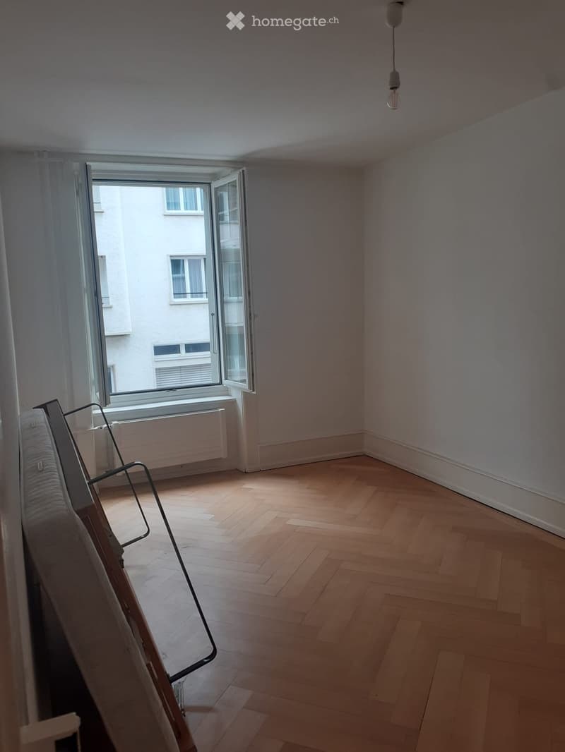 Empty room with wooden flooring, large window with white curtains, and a radiator below the window. No furniture or equipment present.