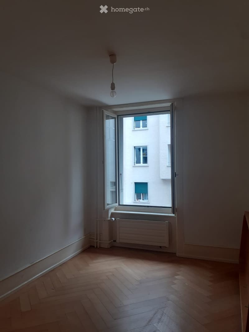 Empty room with wooden flooring, white walls, and an open window showing a view of a neighboring building.