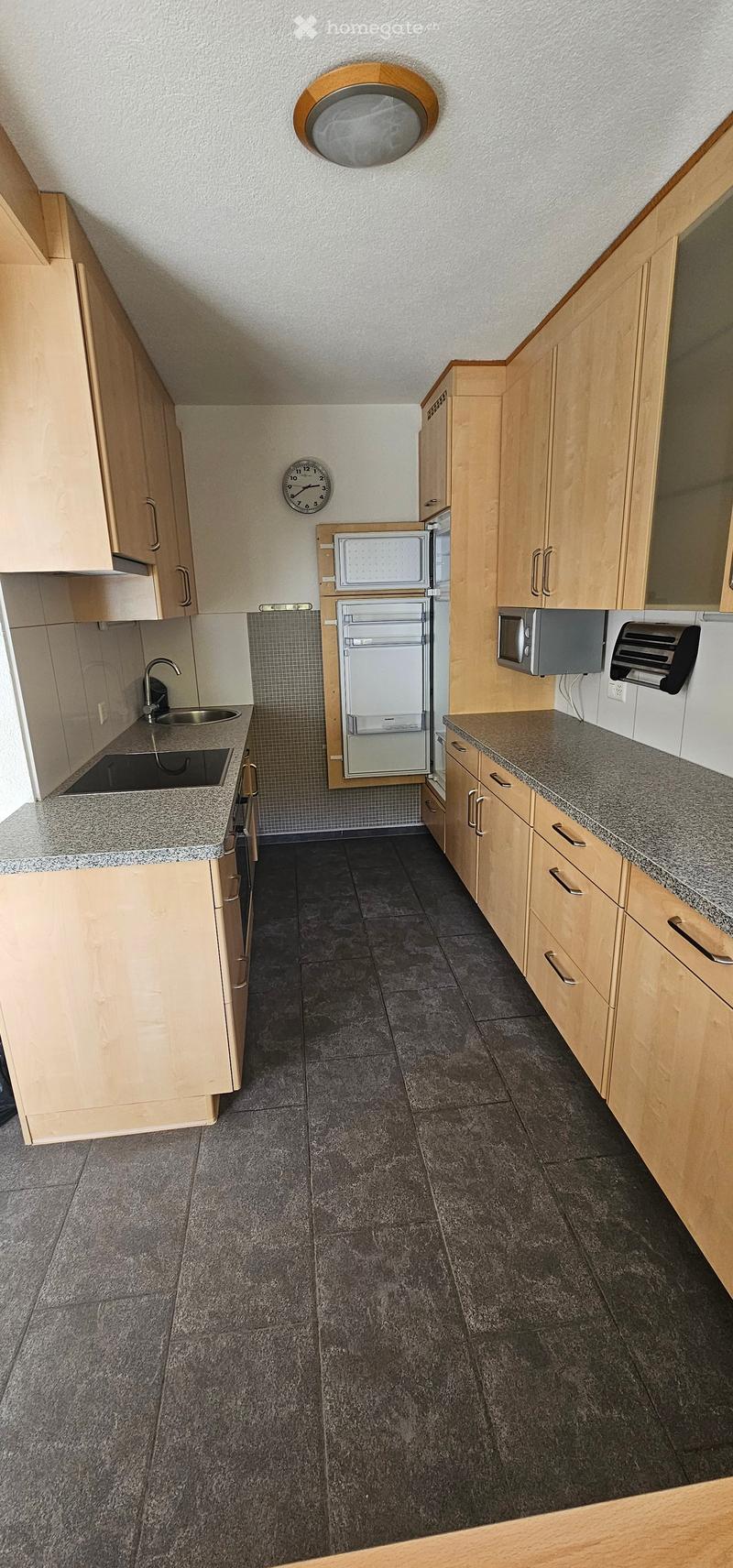 Modern kitchen with light wooden cabinets, granite countertops, dark tiled floor, sink, fridge, microwave, and a clock on the wall.