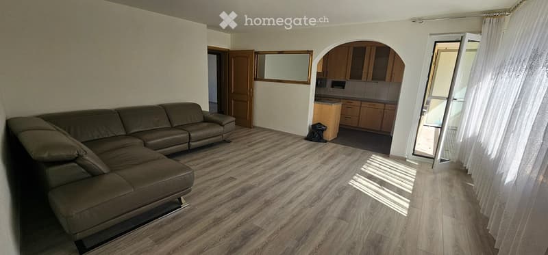 Spacious living room with a large sectional sofa, wooden flooring, and an open kitchen area with cabinets and a countertop.