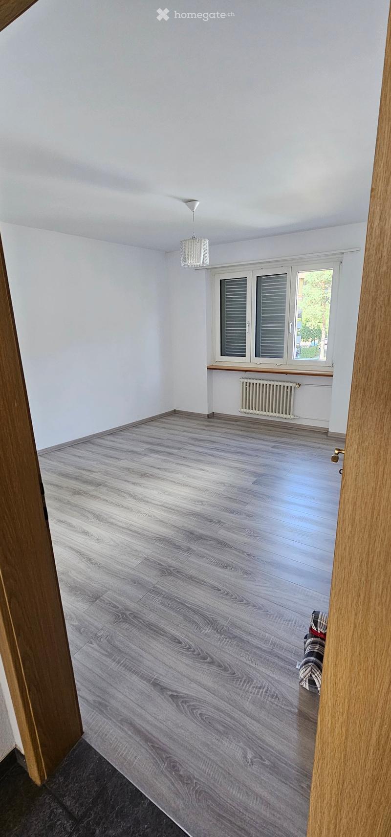 Empty room with light grey wooden flooring, white walls, and a window with shutters. A radiator is visible below the window, and a small piece of furniture or item is partially visible in the corner.