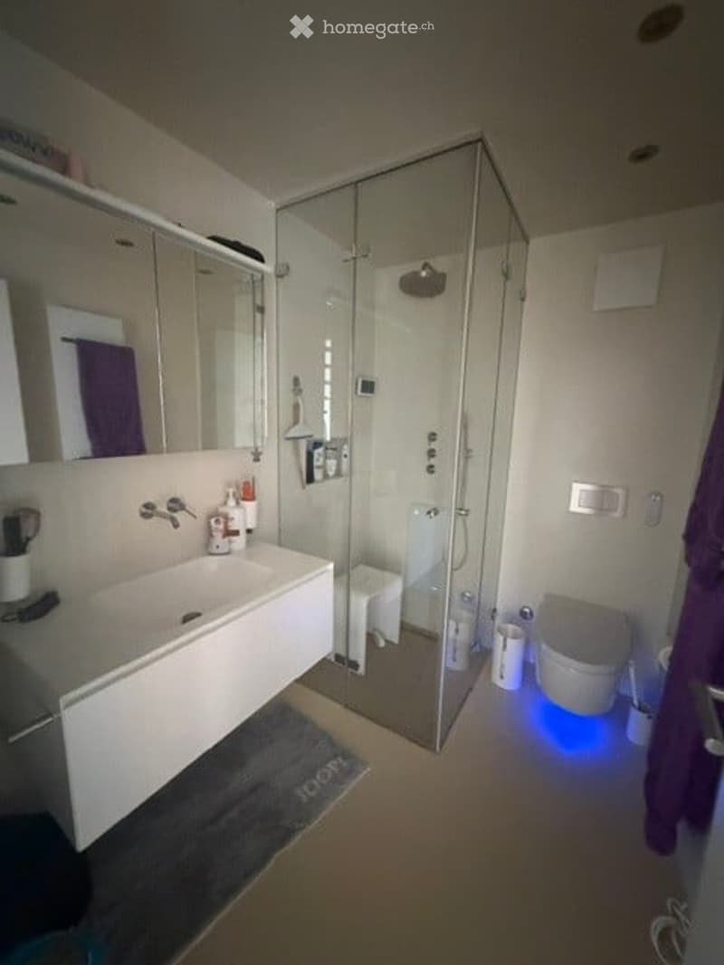 Modern bathroom with a white sink, large mirror, glass-enclosed shower, toilet, and purple towel. Blue LED lighting accents the floor near the shower area.