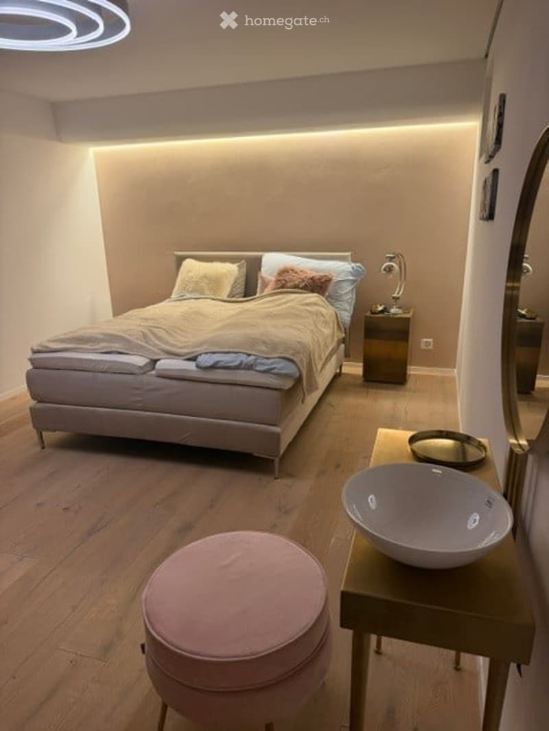 Modern bedroom with a large bed, nightstand with lamp, pink stool, and a small table with a bowl on it.