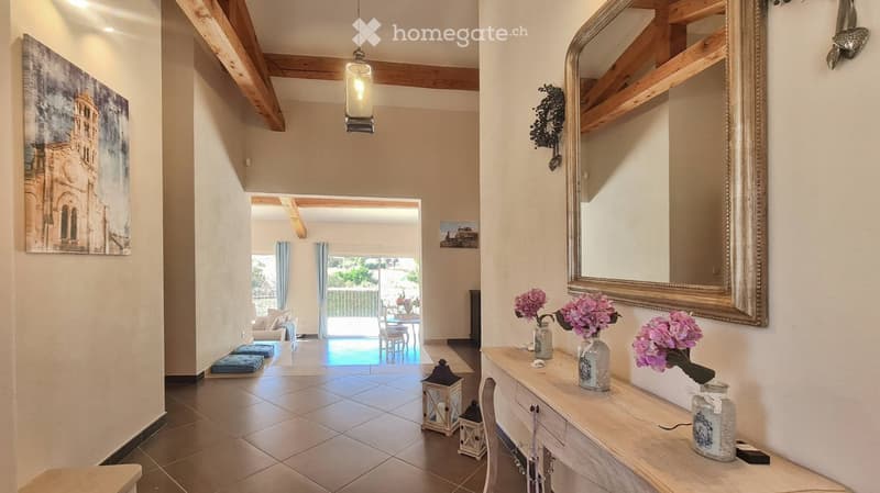 Elegant hallway with exposed wooden beams, a large mirror, decorative vases with pink flowers, and a view to a living area with a sofa and dining table.