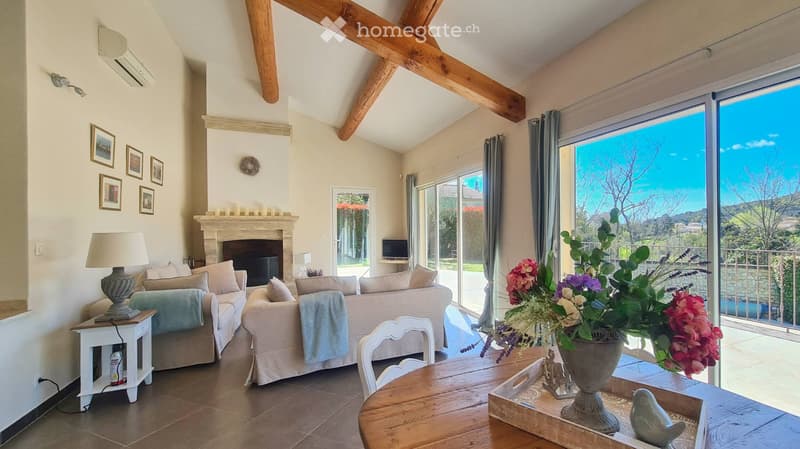 Bright living room with exposed wooden beams, fireplace, large windows, and a view of the landscape. Features a sofa, coffee table, dining table, and decorative items.