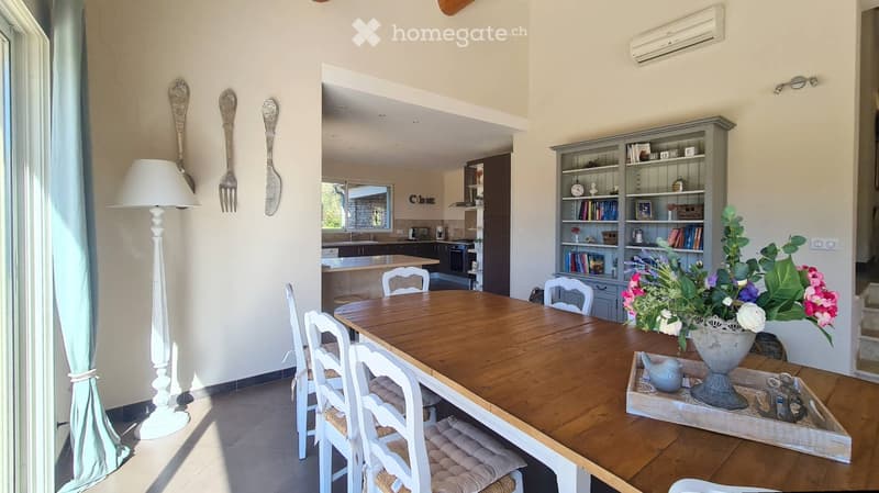 Bright dining area with a wooden table, white chairs, bookshelf, and decorative items. The room features a floor lamp, large windows, and a view into the kitchen area.