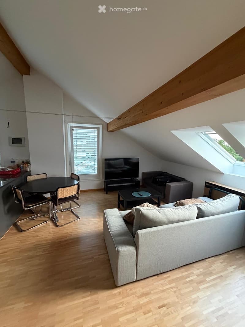 Bright living room with wooden flooring, a grey sofa, coffee table, dining table with chairs, and a television. Skylight windows provide natural light.