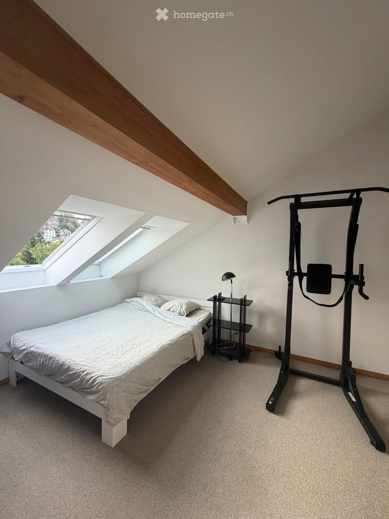 Bedroom with a white bed, nightstand, lamp, and exercise machine. The room has skylight windows and exposed wooden beams on the ceiling.