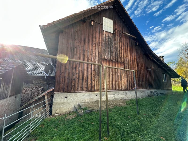 Traditional wooden house with a pitched roof, stone base, and a fenced yard with stacked firewood.