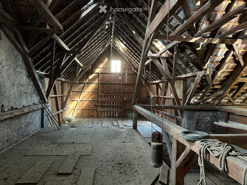An empty attic space with exposed wooden beams, stone walls, and a concrete floor. Natural light streams in from small windows near the roof.
