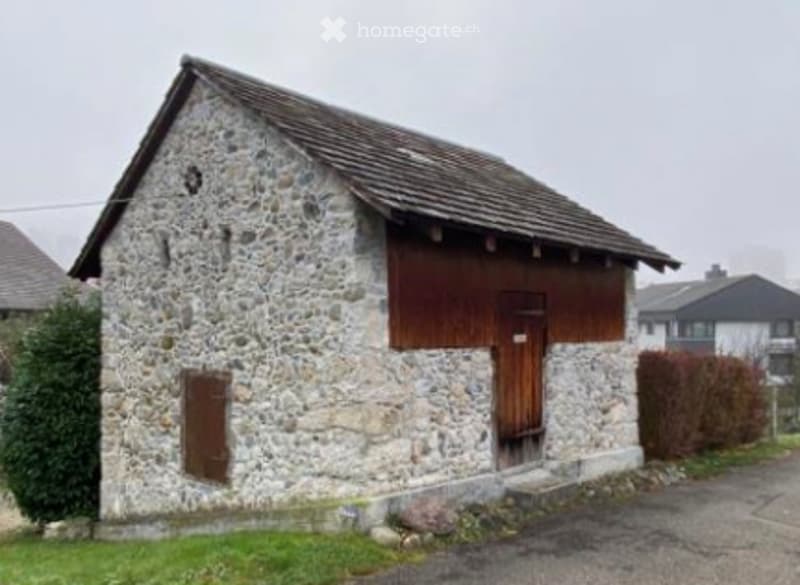 A small stone building with a wooden door and a slanted roof, surrounded by greenery and other structures.