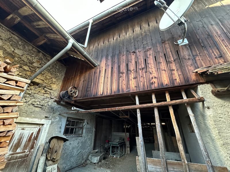 A rustic wooden and stone building with a partially open wooden structure, showing a mix of wooden planks, stone walls, and a satellite dish on the roof.