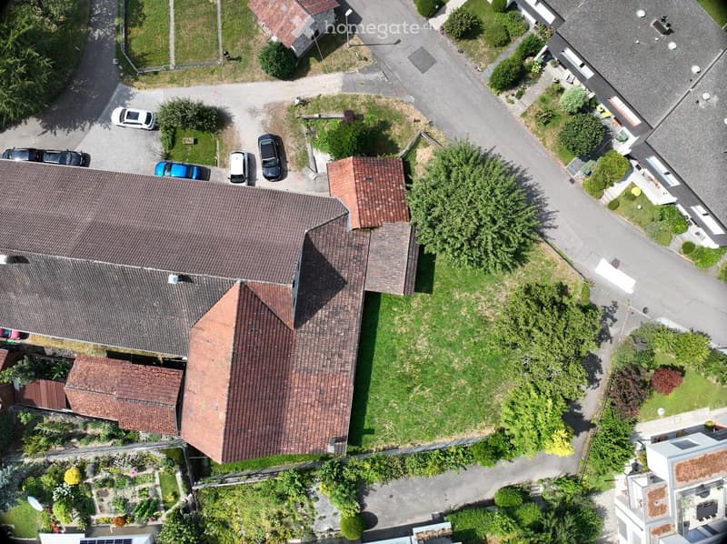 Aerial view of a residential house with a tiled roof, surrounded by a well-maintained garden and driveway with parked cars.