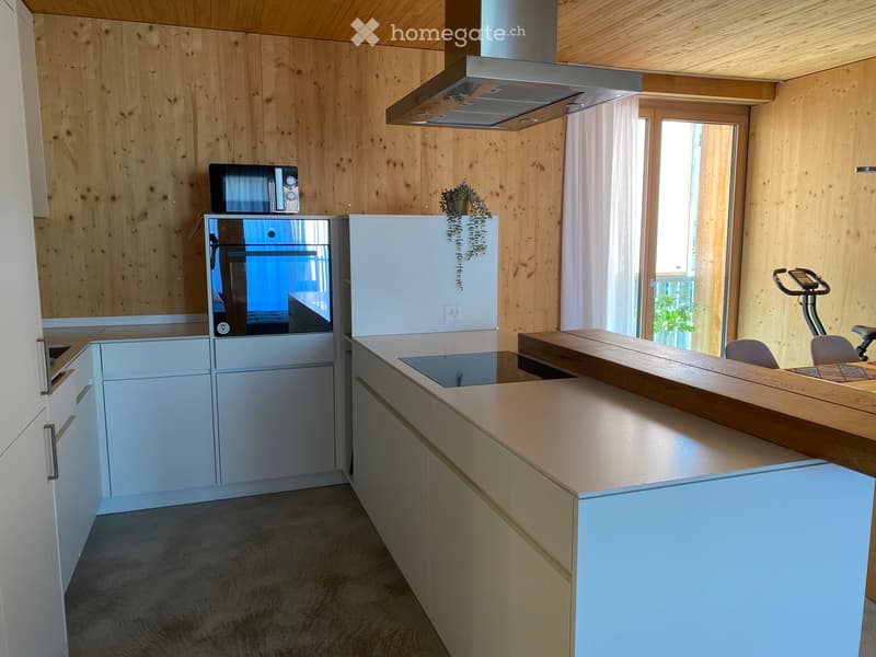 Modern kitchen with light wood walls, white cabinets, stainless steel appliances, and a wooden kitchen island. An exercise bike is positioned near the window.
