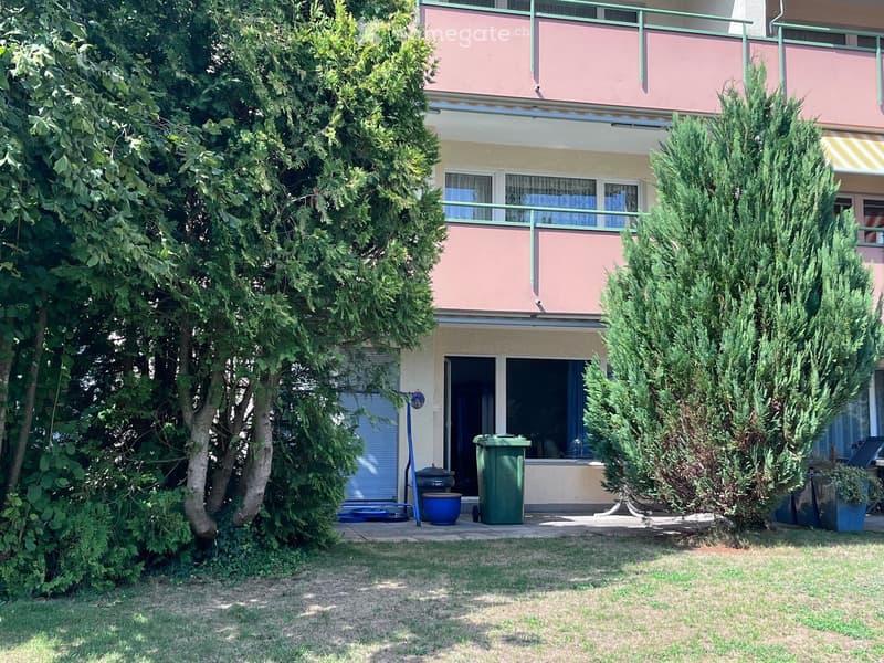 A two-story building with pink and yellow exterior, surrounded by greenery and trees. Features include a balcony, a small patio area with a green trash bin, and a blue barbecue grill.