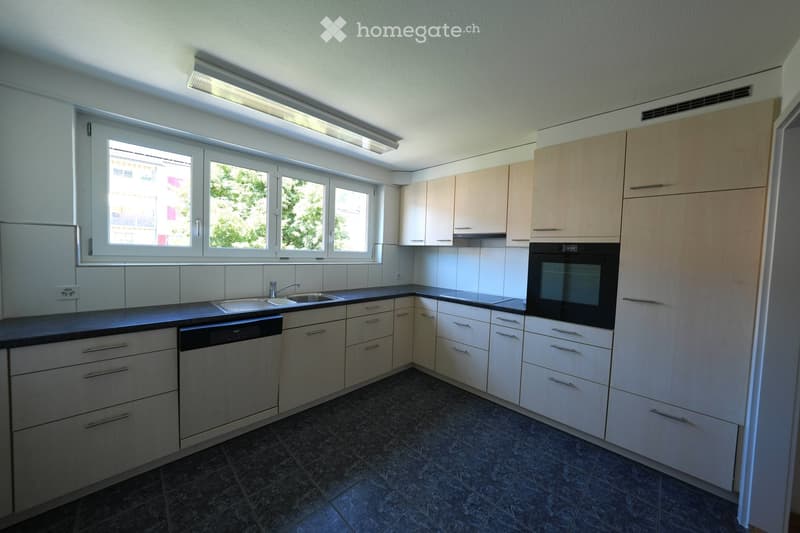 Modern kitchen with white cabinets, dark countertops, and stainless steel appliances including a sink, oven, and dishwasher. Large windows provide natural light.