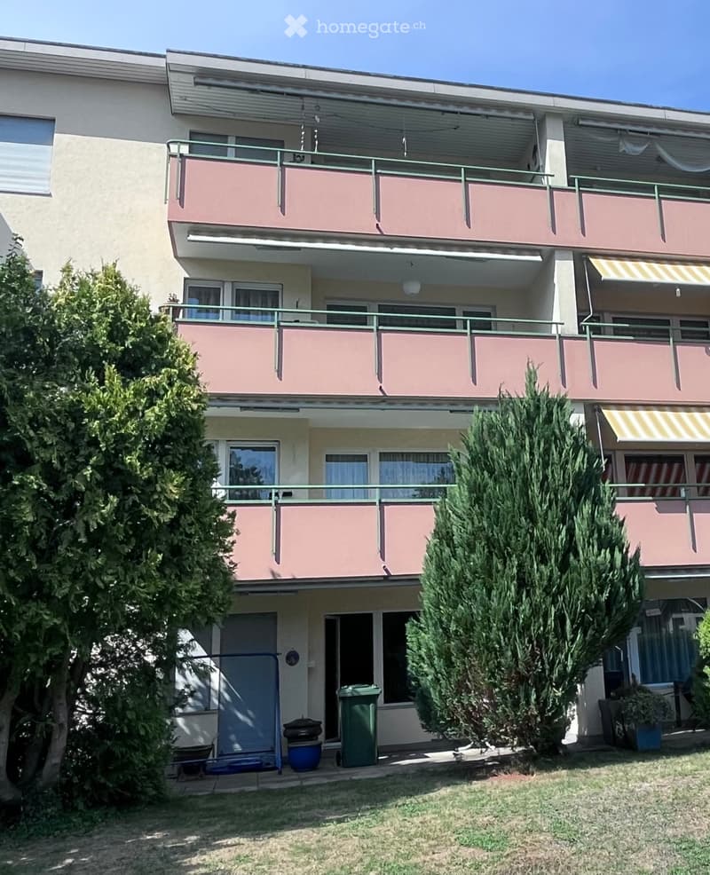 A three-story residential building with balconies, green railings, and multiple windows. The ground floor features a small garden area with potted plants, a trash bin, and a blue container.