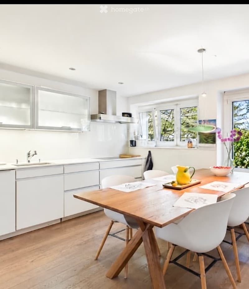 Bright, modern kitchen with white cabinets, a central dining table with chairs, and a vase of flowers on the table. The space features a sink, faucet, and a window with a view of greenery.