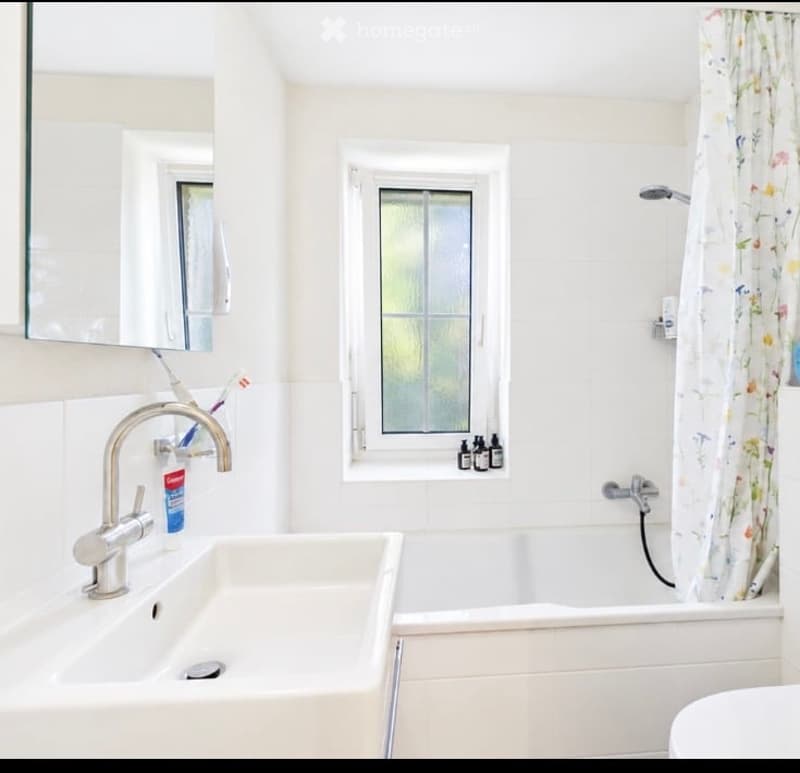 A modern bathroom with a white sink, mirror, bathtub, and shower curtain. The room features a window with frosted glass and a sleek chrome faucet.