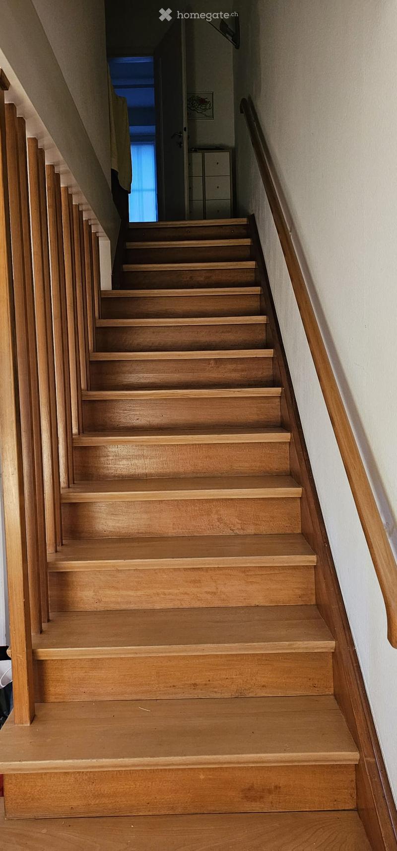Wooden staircase with a handrail, leading upwards, with a small room visible at the top.
