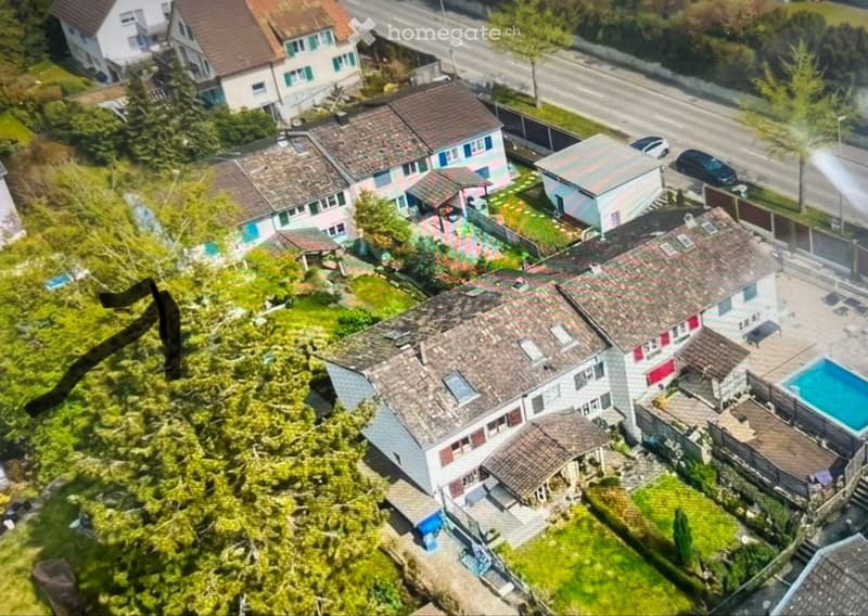 Aerial view of a residential area with multiple houses, gardens, and a swimming pool visible in the foreground.