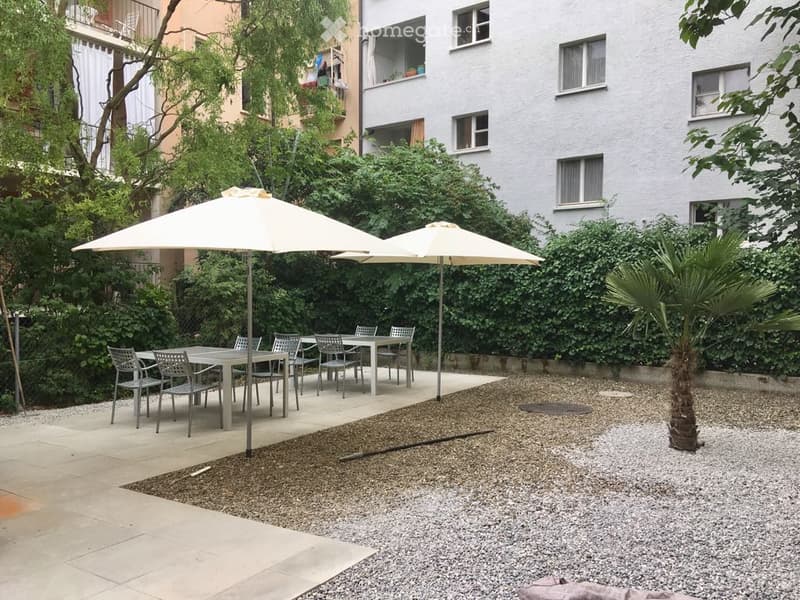 outdoor seating area with tables, chairs, umbrellas, gravel path, greenery, building