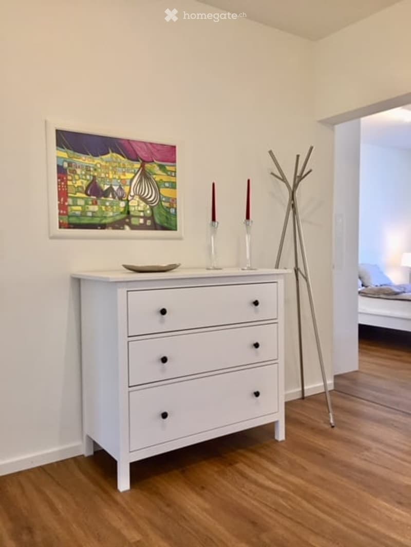 White wooden floor, white walls, a white dresser with four drawers, a plate on top, two candlesticks with red candles, a silver coat stand, a framed painting on the wall, a glimpse of a bed and pillow in the adjacent room.