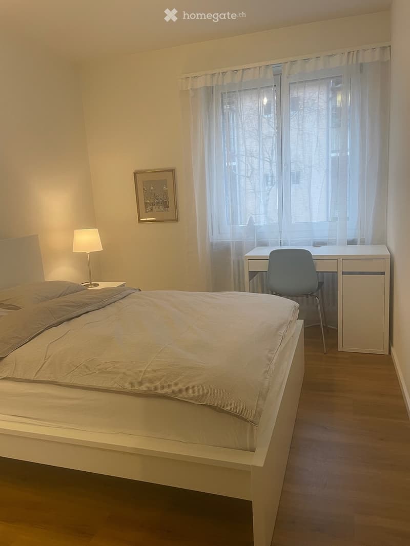 Bed with white bedspread, 2 pillows, bedside table with a lamp, desk with chair, picture frame on the wall, window with curtains