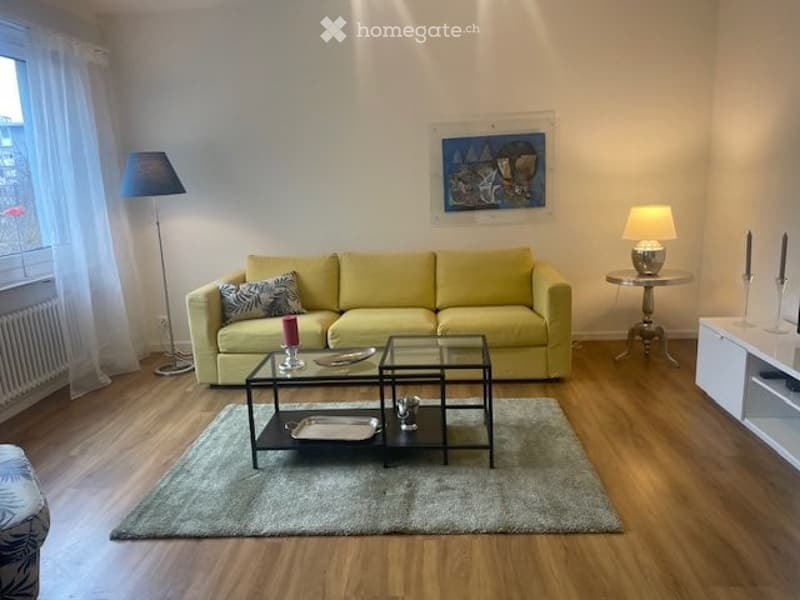 Bright and spacious living room with a large yellow sofa, glass coffee table, and a decorative painting on the wall. The room has hardwood floors and a rug, and appears to have access to a balcony or terrace through the window.