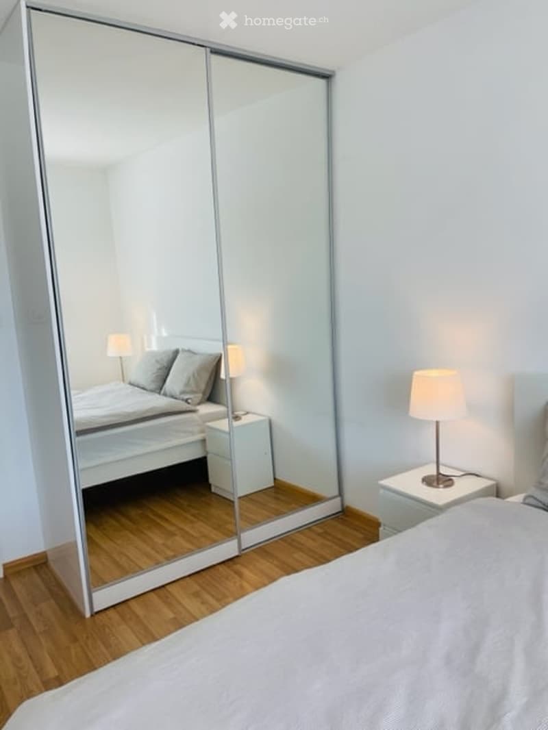 Bedroom with white walls, large sliding mirror, white bed, white nightstands with lamps, white linens, and hardwood floors.
