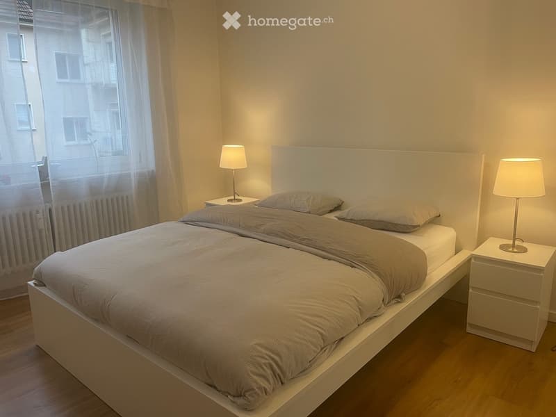 a spacious bedroom with a white bed, two lamps, white nightstands, and a white headboard, with a large window covered by white curtains and white walls, offering a serene view outside.