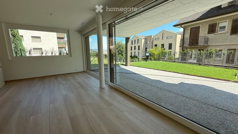 Empty room with wooden flooring, large glass sliding doors leading to a patio, and a view of a green lawn and neighboring buildings.