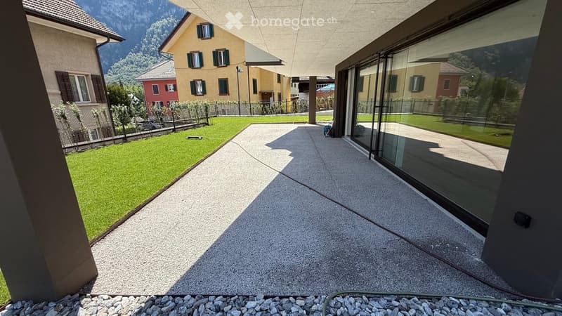 Modern outdoor patio with concrete flooring, large glass windows, and a view of a green lawn and surrounding houses.