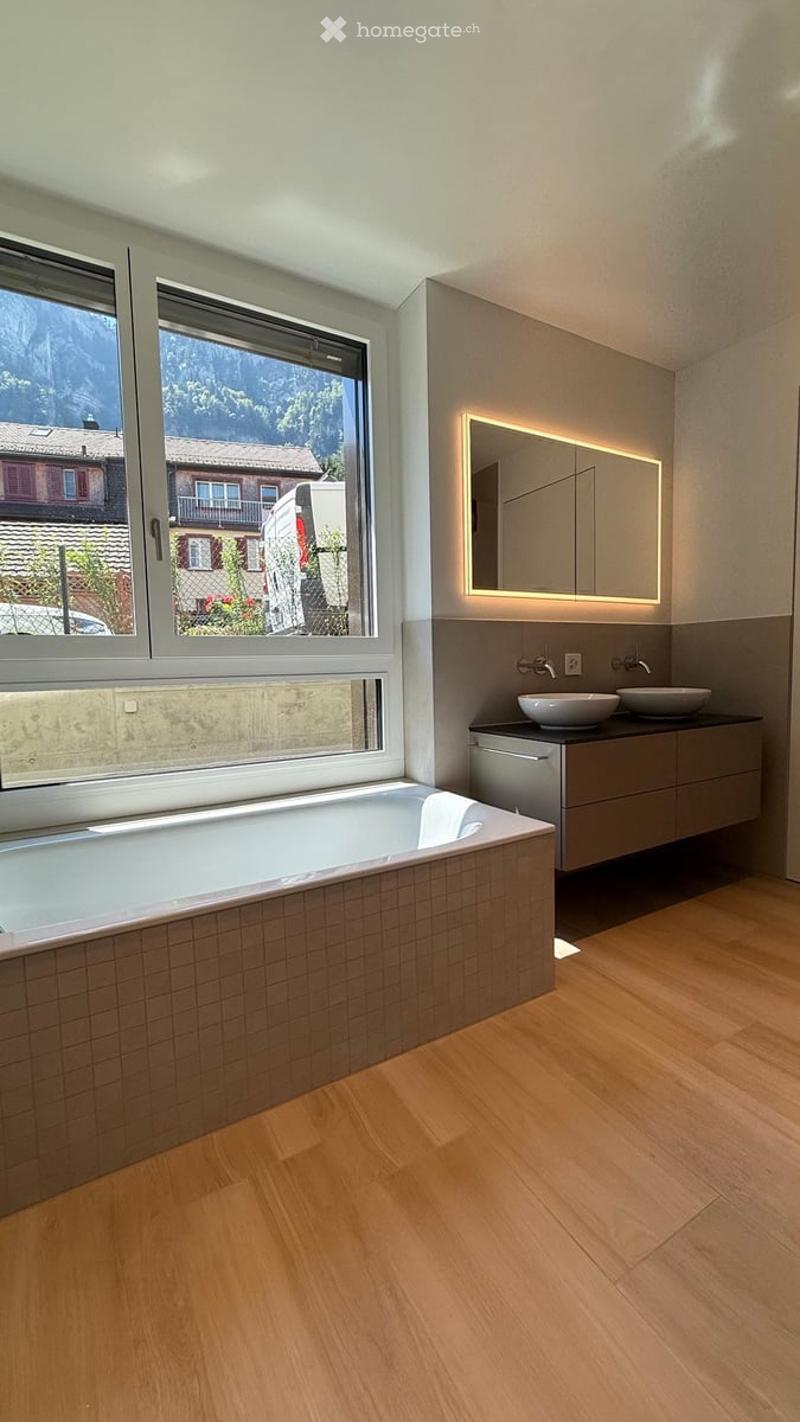 Modern bathroom with a large window offering a scenic view, featuring a bathtub, double sink, mirrored cabinet, and wooden flooring.