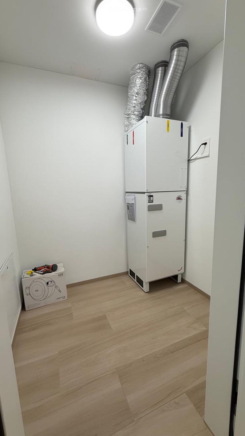 Empty room with light wooden flooring, white walls, and a large white appliance unit with ventilation pipes. A small toolbox and a vacuum cleaner box are placed on the floor.