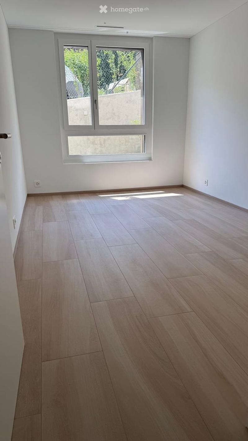 Empty room with light wooden flooring and white walls, featuring a large window with a view of greenery outside.