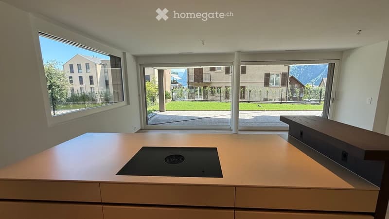 Modern kitchen with an induction cooktop, a wooden bench, large windows, and a view of the outdoor area and surrounding buildings.