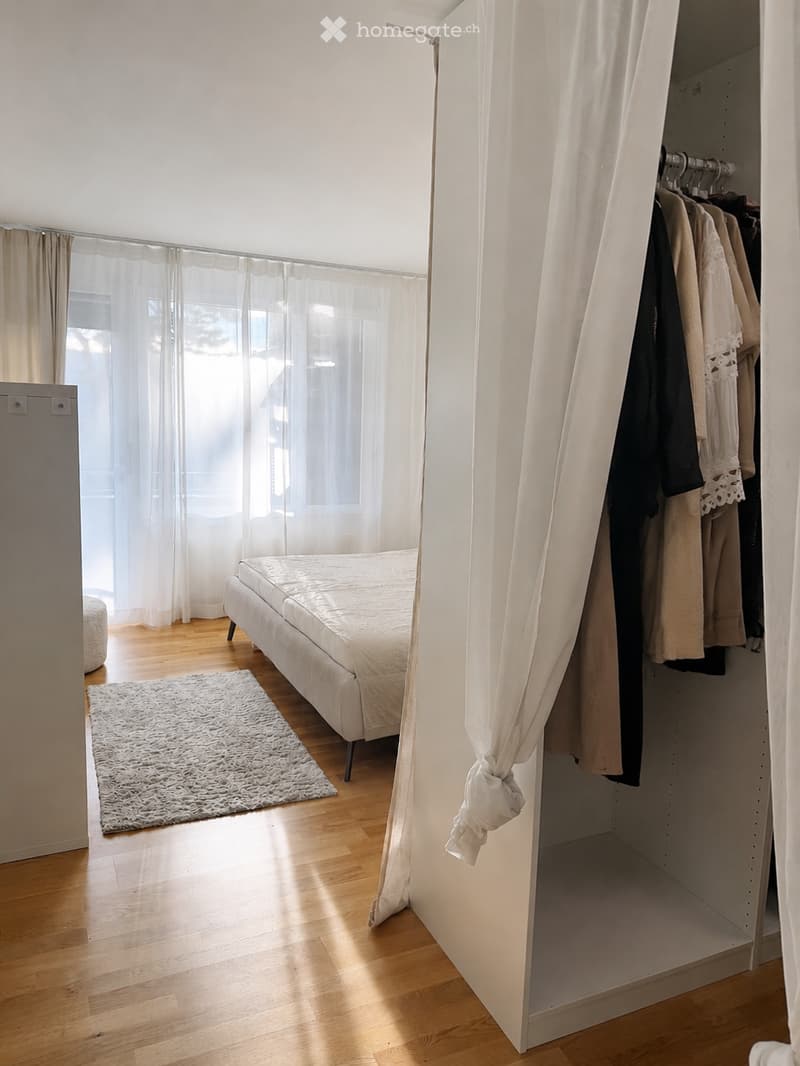 Bright bedroom with wooden flooring, a white bed, a shaggy rug, and an open closet with hanging clothes.