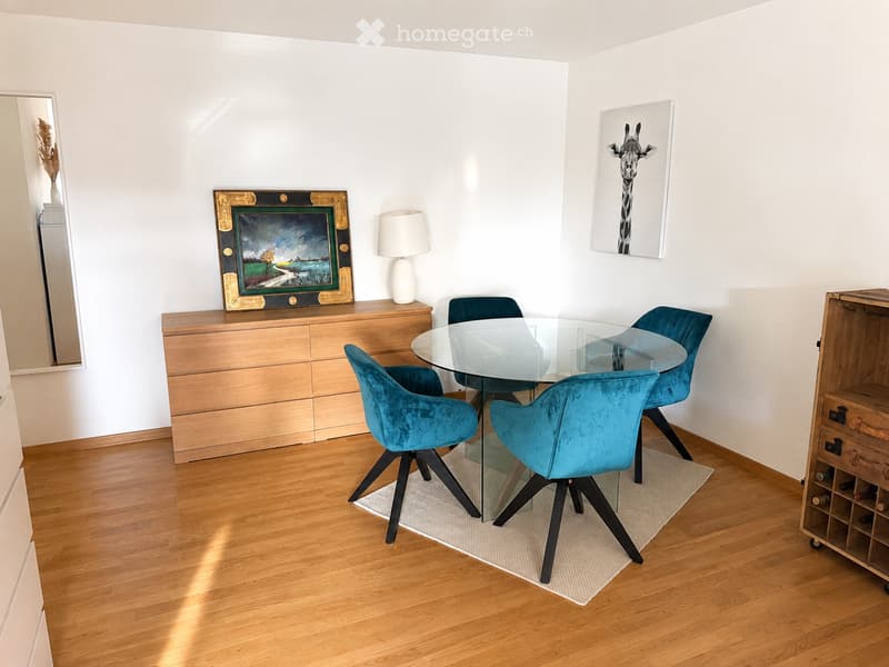 Dining room with a glass table surrounded by four teal upholstered chairs, a wooden sideboard with a lamp and artwork, and a wine rack in the corner.