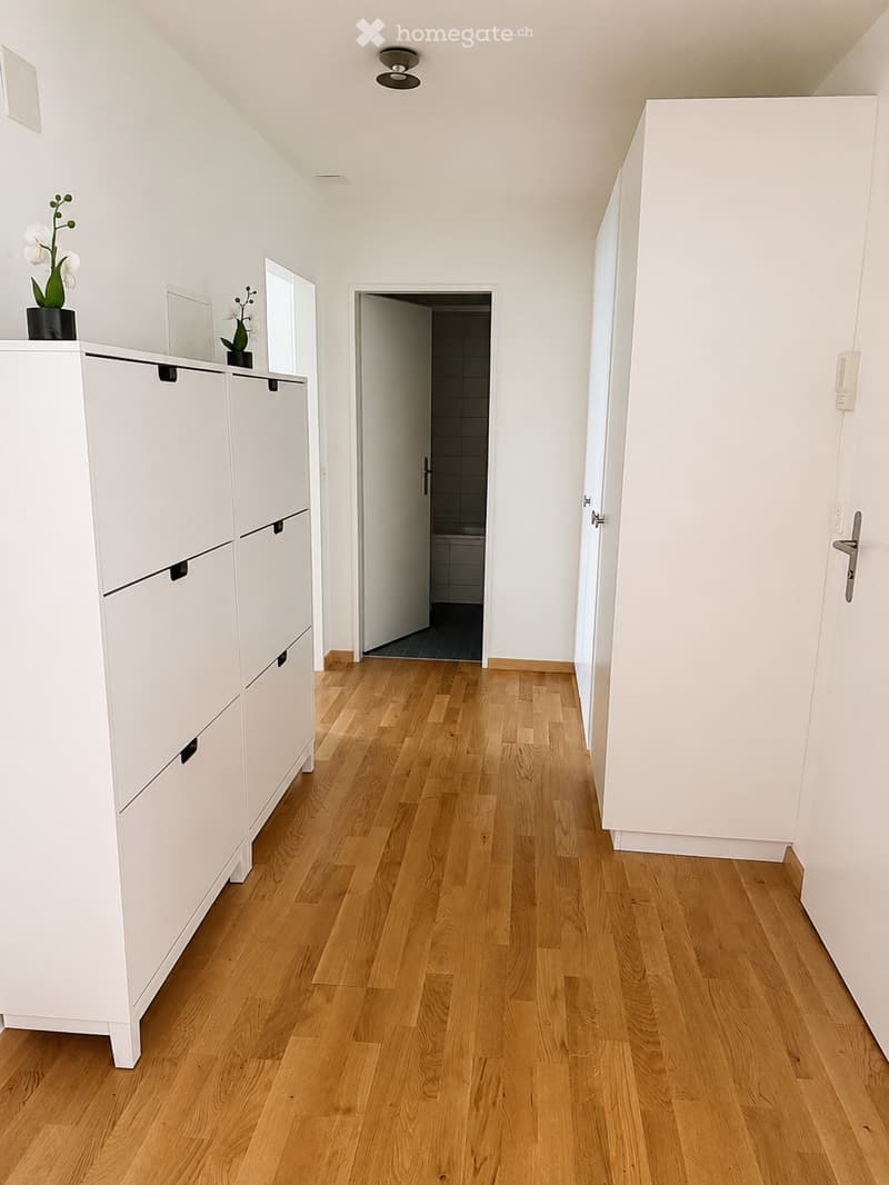 Bright hallway with light wooden flooring, white walls, and a built-in white storage unit with drawers and shelves. A small vase with white flowers is placed on top of the unit. An open doorway leads to a bathroom with tiled walls and a visible bathtub.