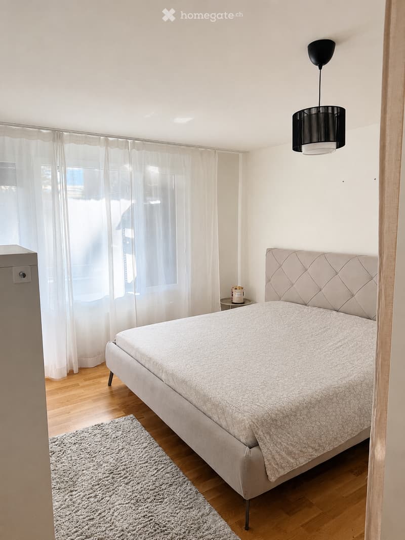 Modern bedroom with a large bed, patterned bedding, a grey shaggy rug, a side table with a lamp, and sheer white curtains covering a large window.