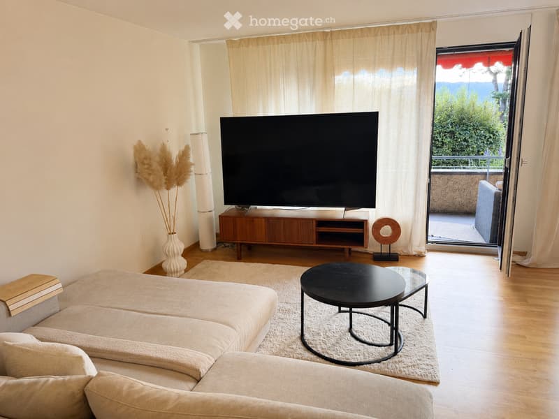 Modern living room with a large flat-screen TV on a wooden stand, a beige sectional sofa, a black coffee table, a glass side table, a decorative vase with dried plants, and a floor lamp. The room features light wooden flooring and large windows with sheer