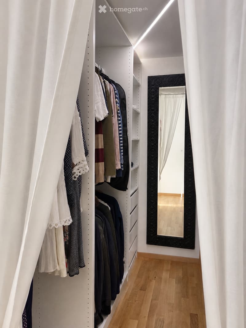 Well-organized walk-in closet with white built-in wardrobe, multiple clothing items, and a large mirror with black frame.