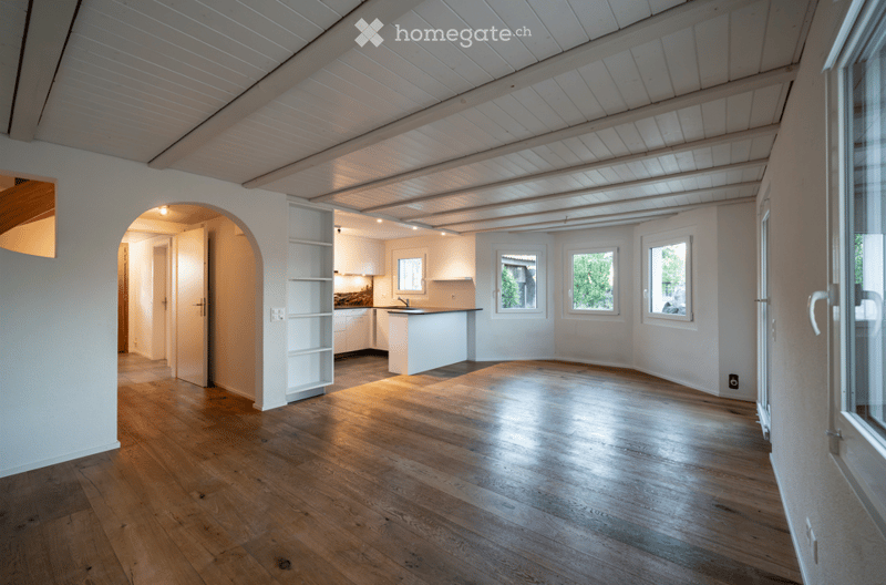 Bright, open living space with wooden flooring, white walls, and large windows. The room features an arched doorway leading to another area and a visible kitchen area with cabinets and a countertop.