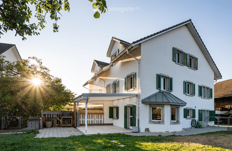 Two-story white house with green shutters, a covered patio area, and a small garden with a grill and seating area.