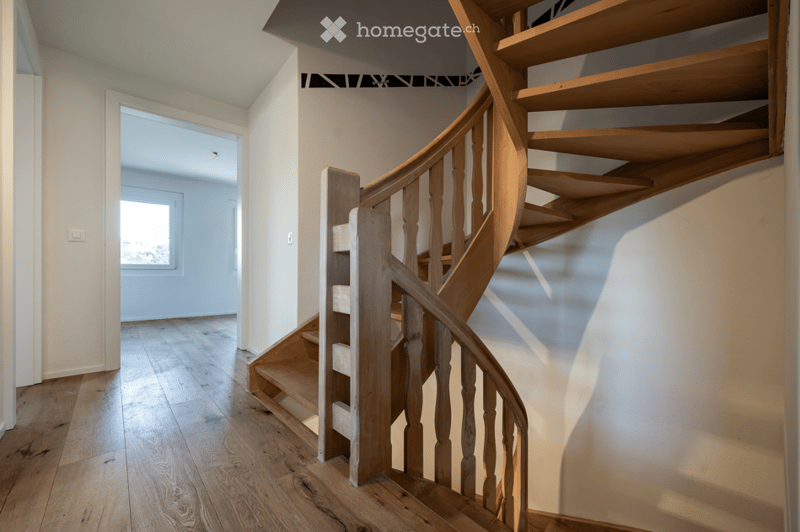 Spacious hallway with wooden flooring and a wooden spiral staircase leading upwards. The walls are painted in a light neutral tone, and there is a visible doorway leading to another room with natural light coming through a window.
