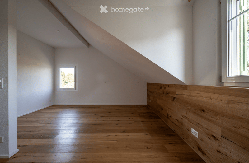 Empty room with wooden flooring, white walls, and two windows allowing natural light. The ceiling has a sloped design typical of an attic or loft space.