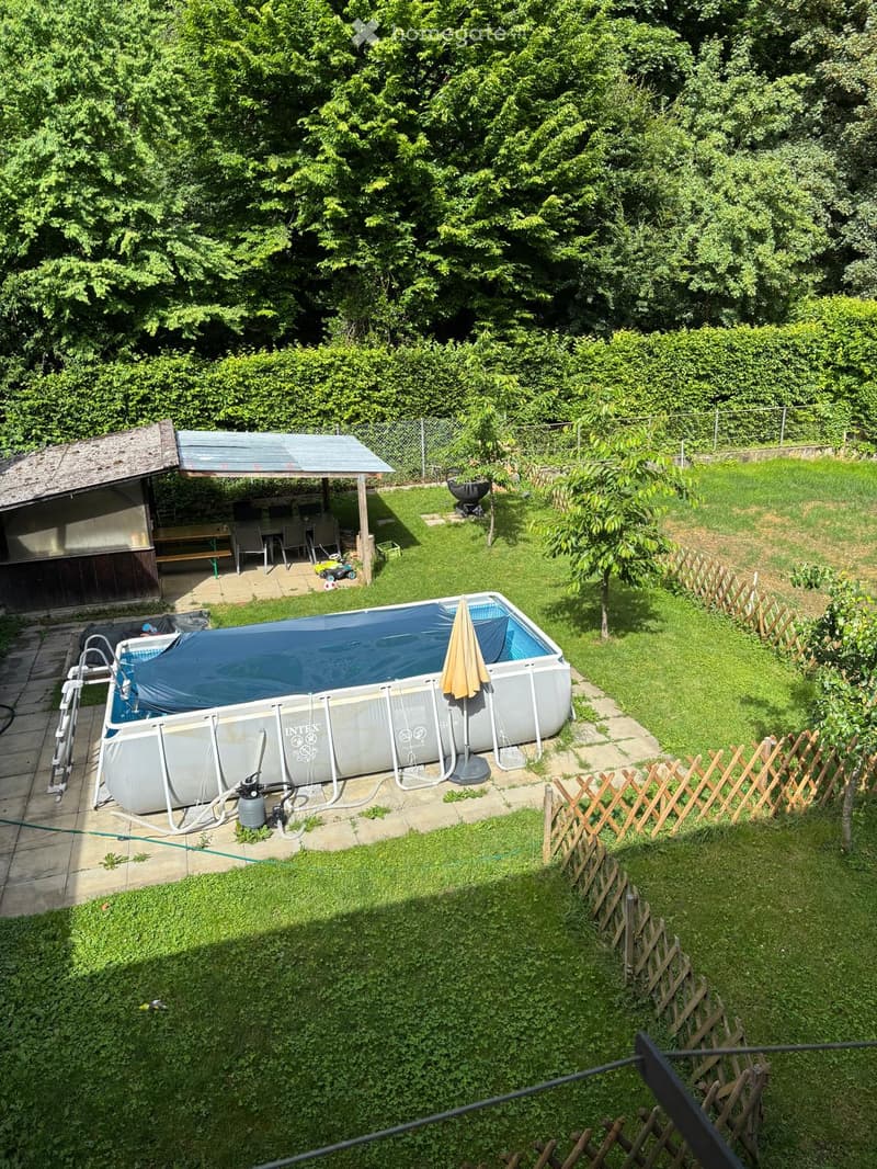 Garden with a covered swimming pool, patio area with chairs and a table, barbecue, and surrounding greenery.
