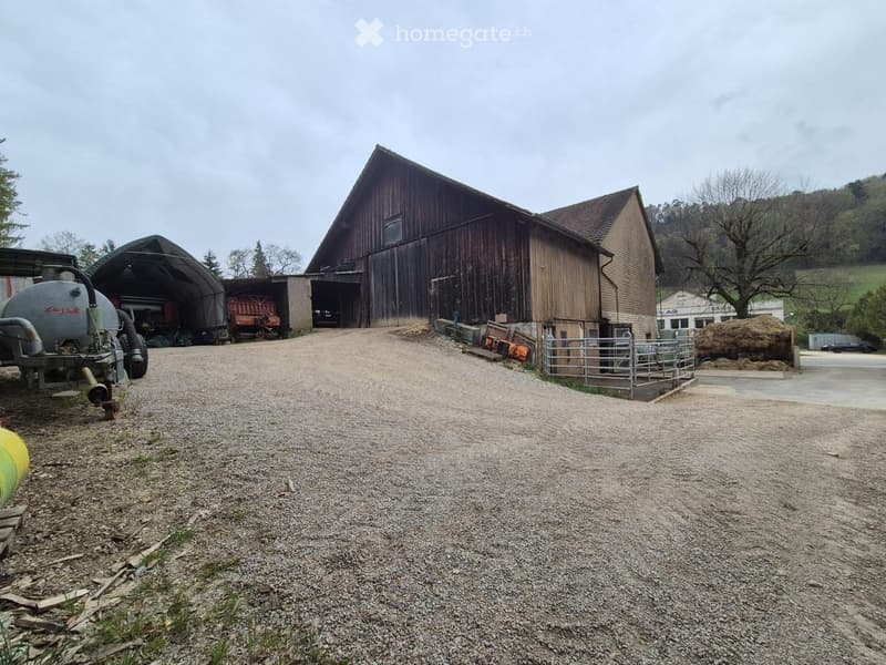 A large wooden barn with a gravel driveway, surrounded by agricultural equipment and a fenced area.