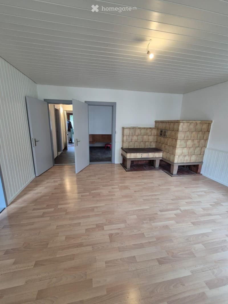 Empty room with light wooden flooring, white paneled walls, and a built-in stone fireplace with a bench. Two open doors lead to adjacent spaces.